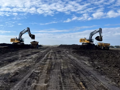 Two excavators loading soil into dump trucks on a large construction site under a partly cloudy sky.