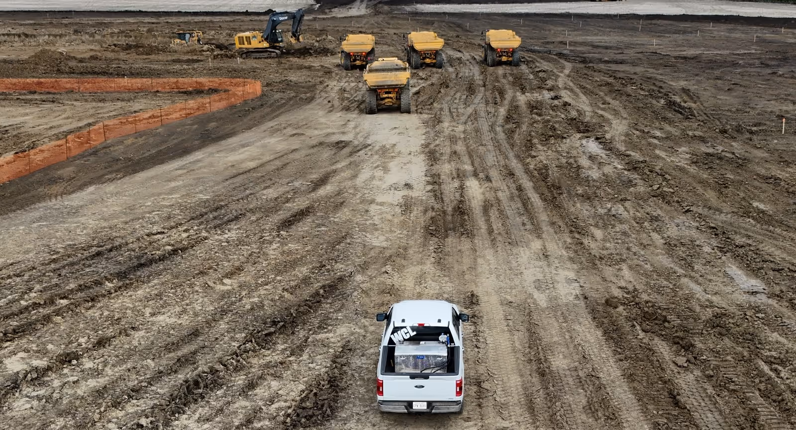 Overhead view of a white pickup truck on a muddy construction site with four yellow dump trucks and an excavator in the background.