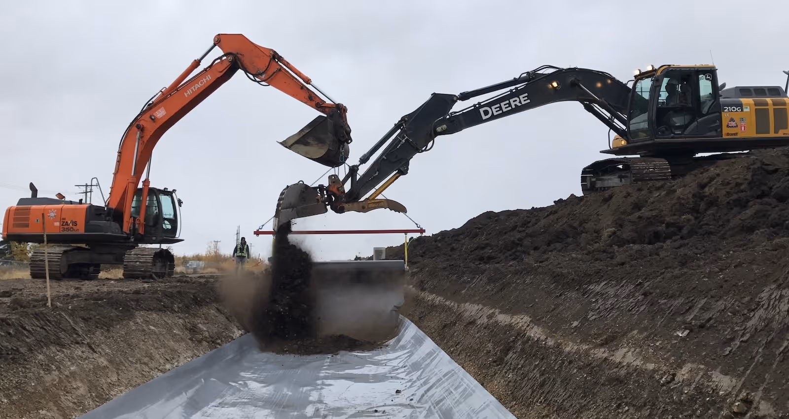 Two excavators, one orange and one black, working together to dump soil into a trench lined with a plastic sheet.