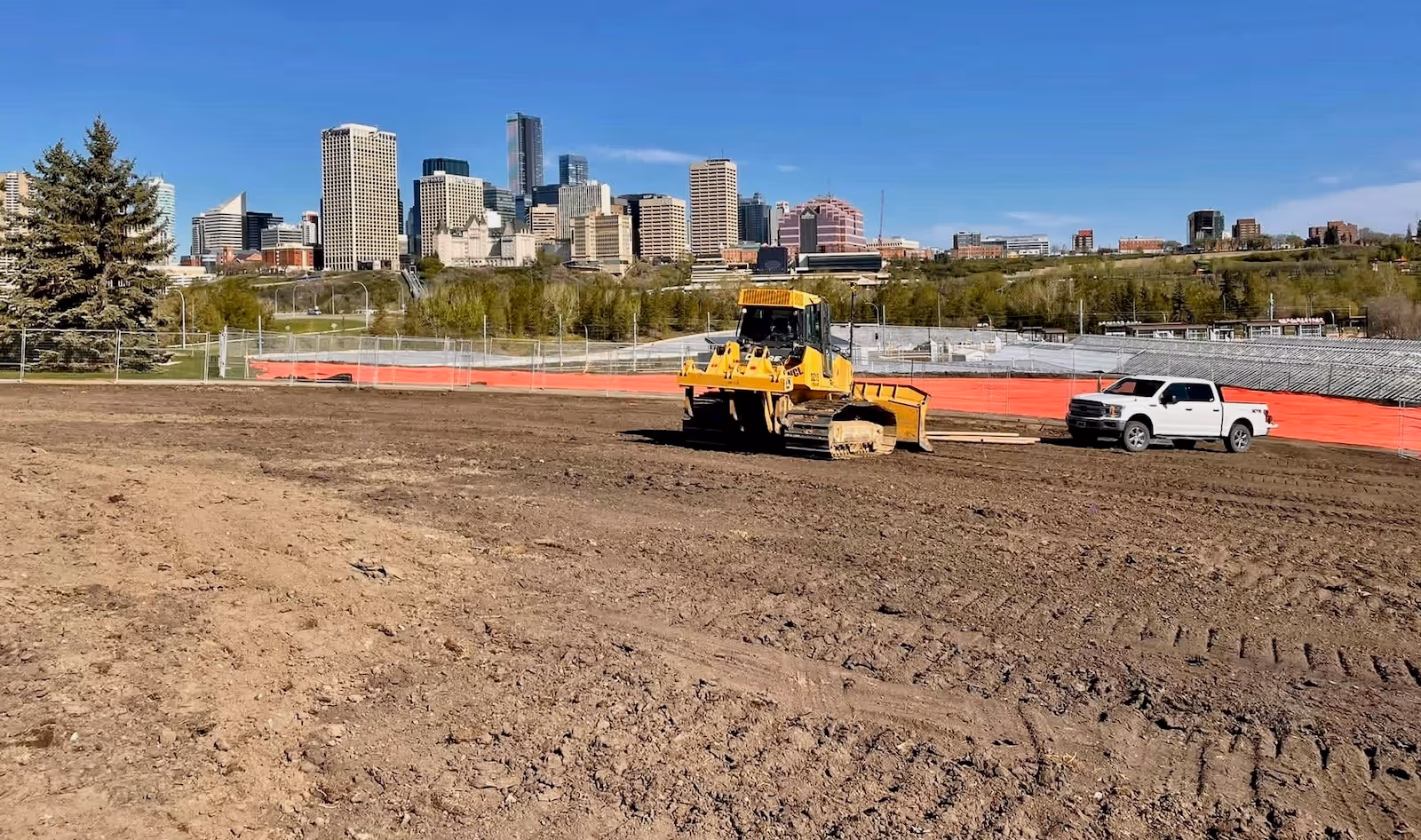 Construction site with a yellow bulldozer and a white pickup truck against a city skyline under a clear blue sky.