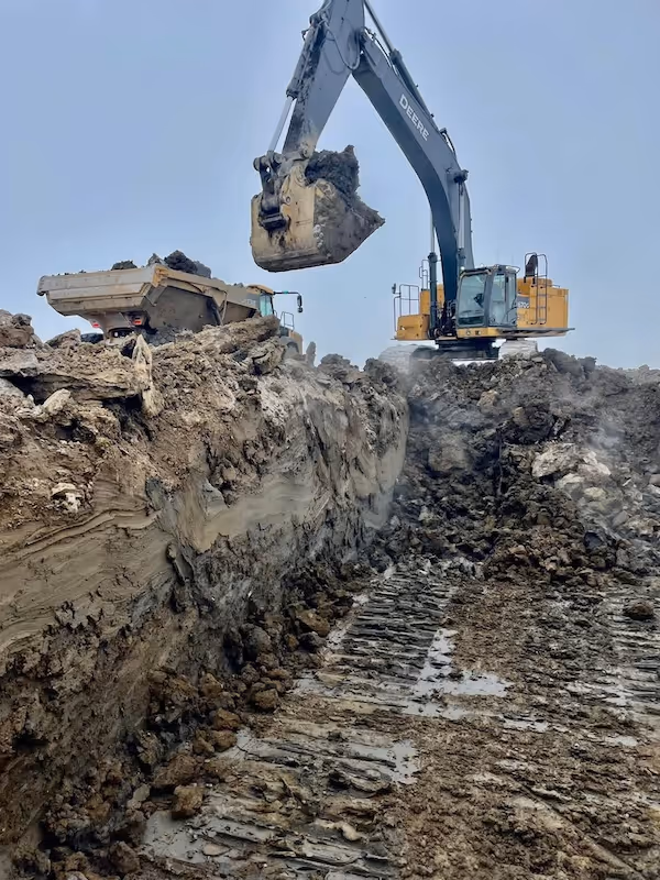 Large excavator loading dirt into a dump truck at a muddy construction site under a clear sky.