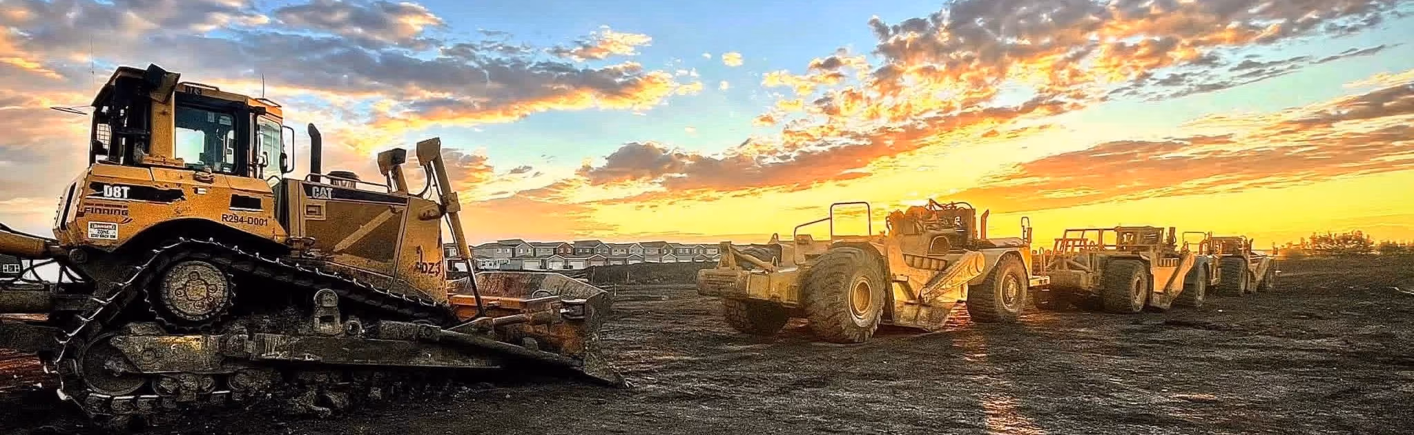 Yellow construction vehicles lined up on dirt at a site during a colorful sunset with houses in the background.
