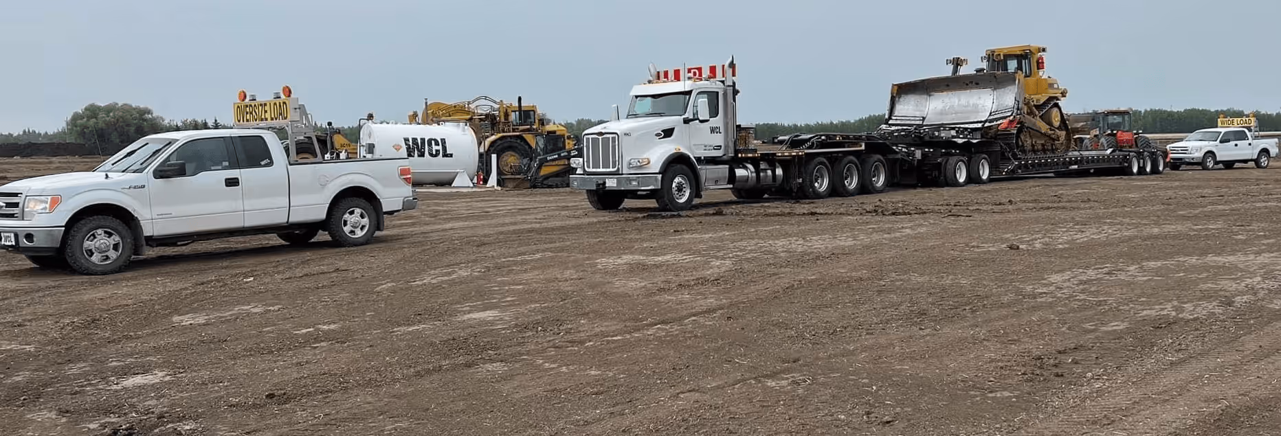 Heavy-duty truck transporting a large bulldozer with white escort pickup trucks labeled 'OVERSIZE LOAD' and 'WIDE LOAD' on a dirt field.