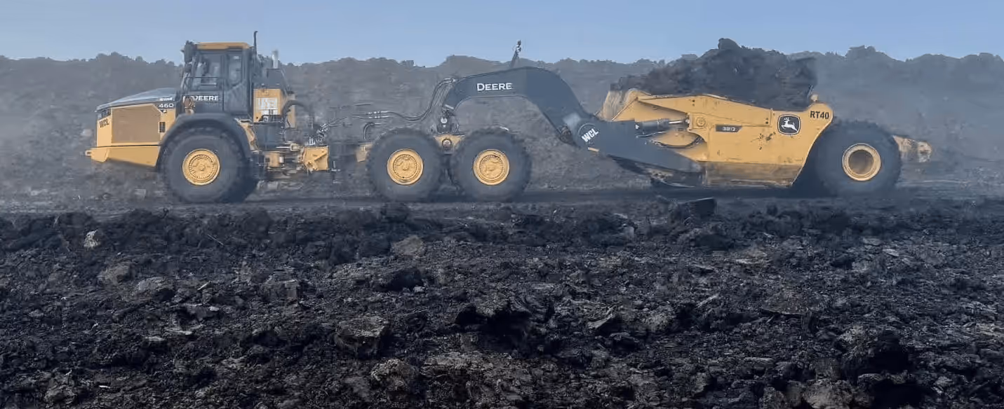 Yellow Deere heavy machinery working on dark soil or coal terrain under clear blue sky.