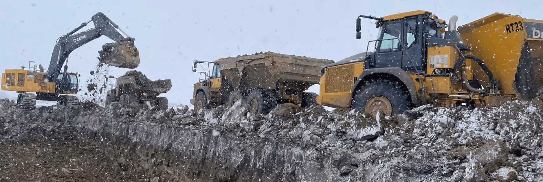 Construction site with a John Deere excavator loading dirt into a dump truck amidst falling snow.