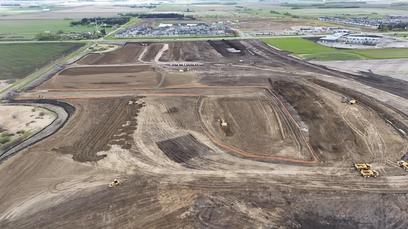 Aerial view of a construction site with earthmoving vehicles on cleared land near residential area and fields.