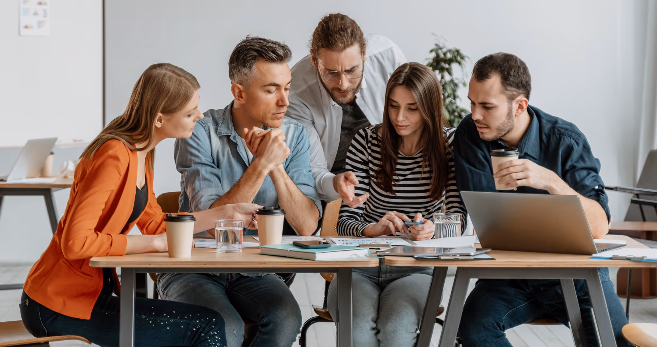 Group of five young professionals collaborating around a table with laptops, documents, and coffee cups in an office.