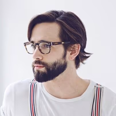 Young man with dark hair, beard, and tortoiseshell glasses wearing a white shirt with striped suspenders looking to the side.