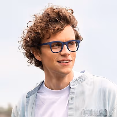 Smiling man with curly hair wearing blue glasses, a white t-shirt, and a light denim shirt outdoors.