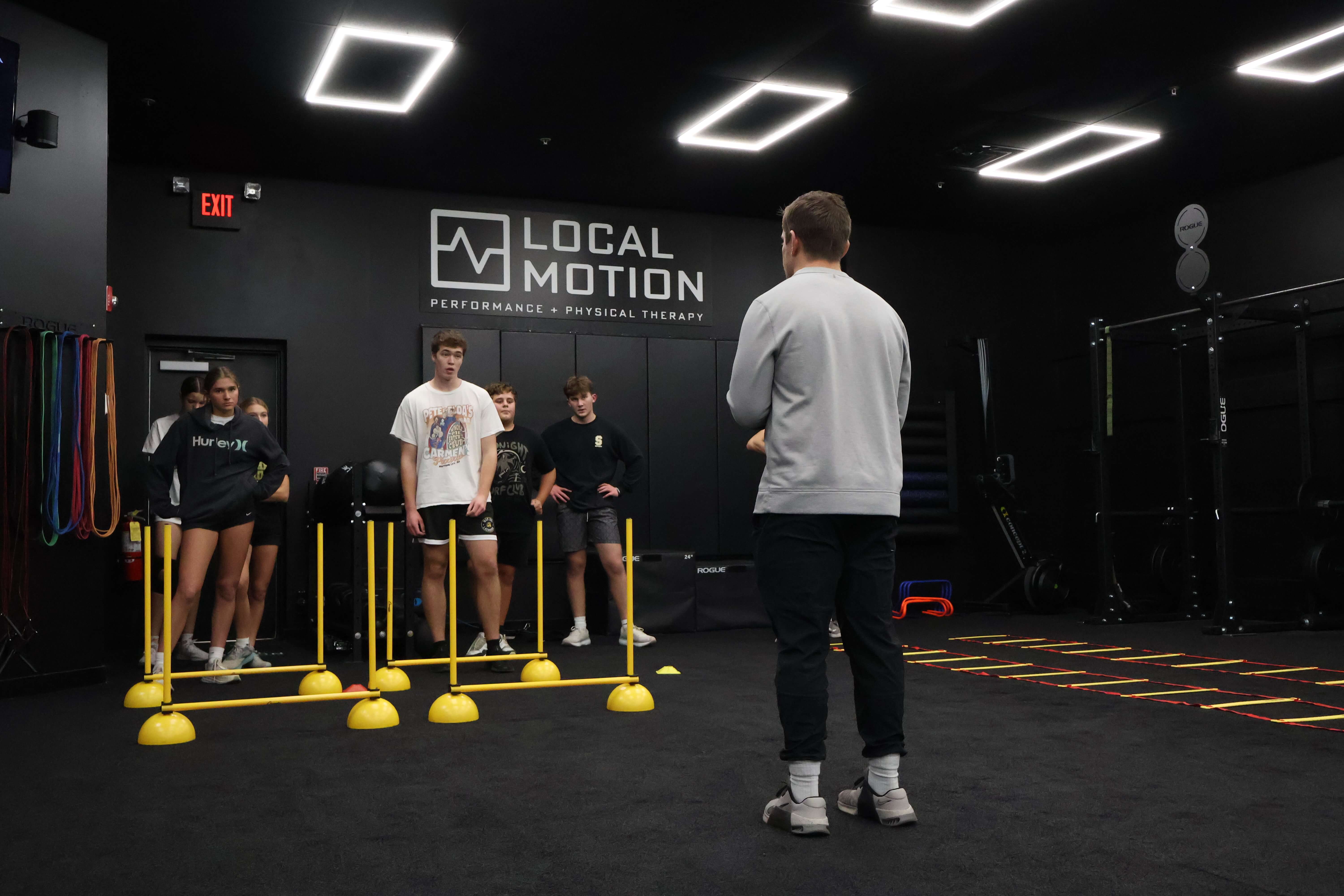Group of young people participating in a fitness training session inside a gym called Local Motion with yellow agility hurdles and ladder drills on the floor.