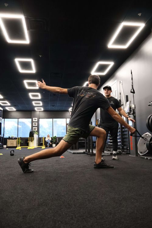 A man in athletic wear stretching in a gym while a trainer watches and instructs him.