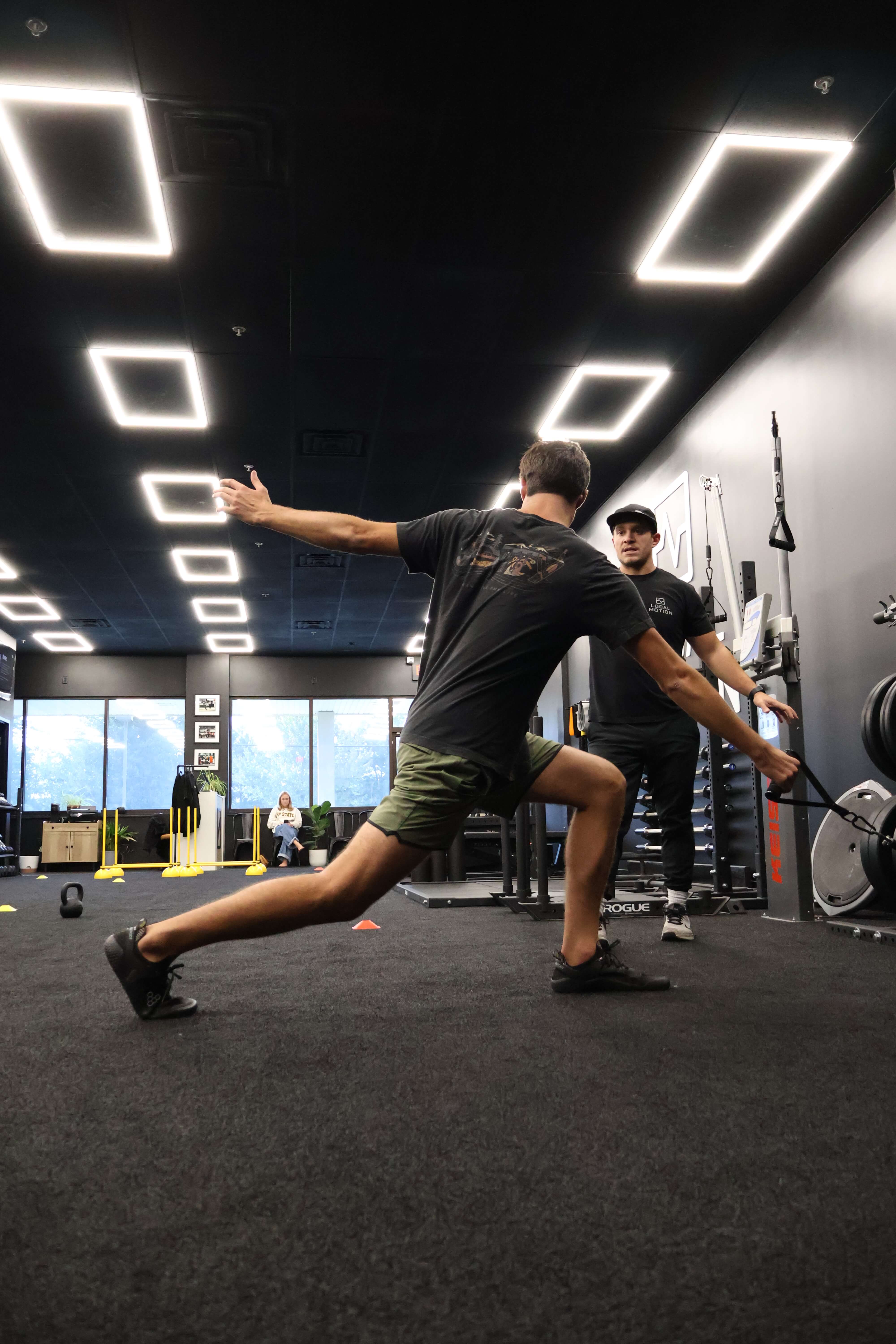 A man in athletic wear stretching in a gym while a trainer watches and instructs him.