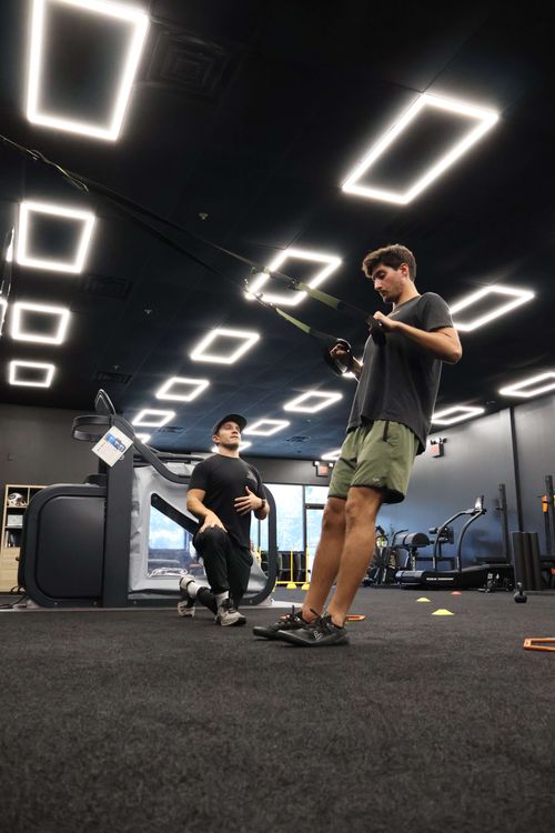 A man in green shorts performing a suspension training exercise while another man kneels nearby coaching him inside a gym.