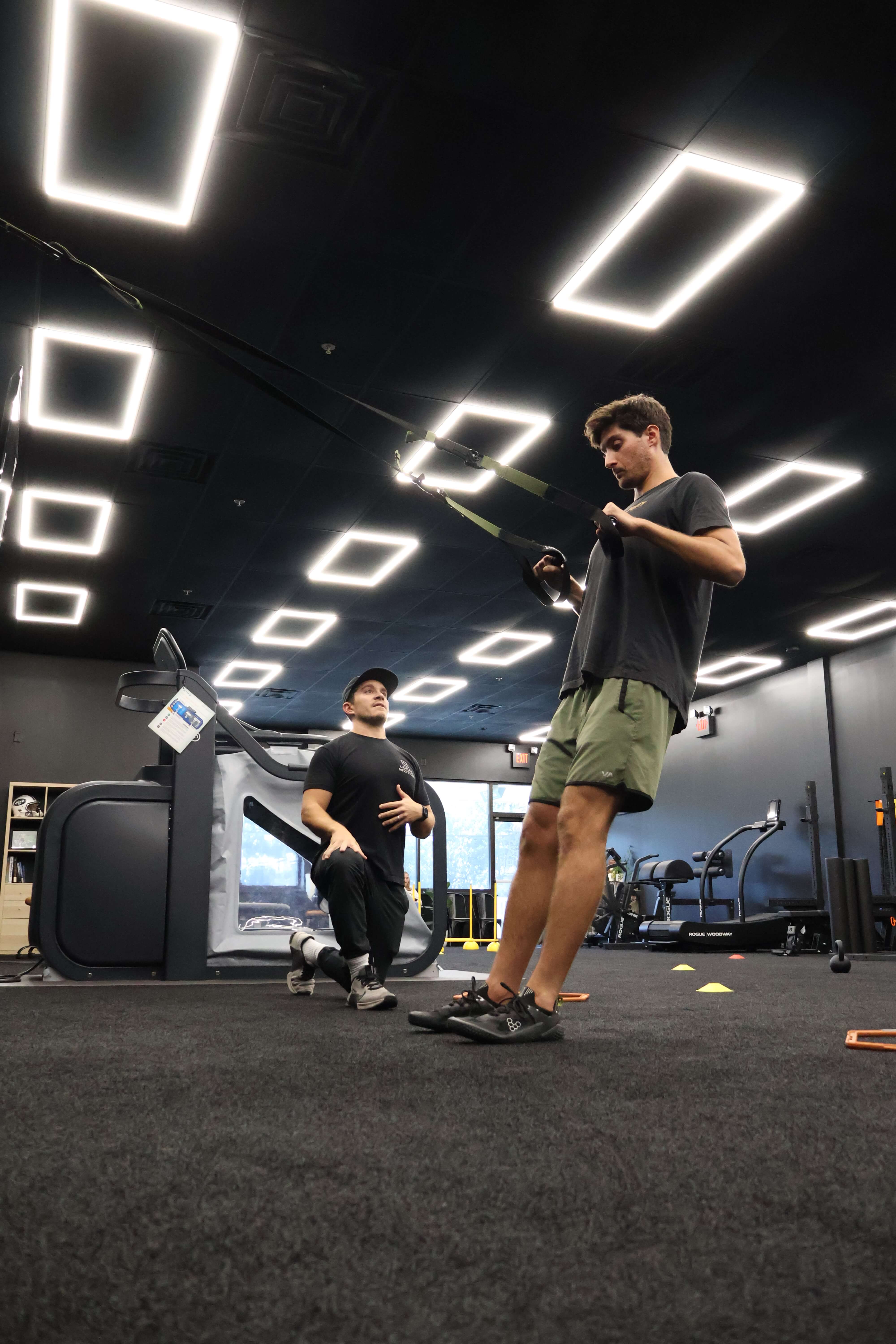 A man in green shorts performing a suspension training exercise while another man kneels nearby coaching him inside a gym.