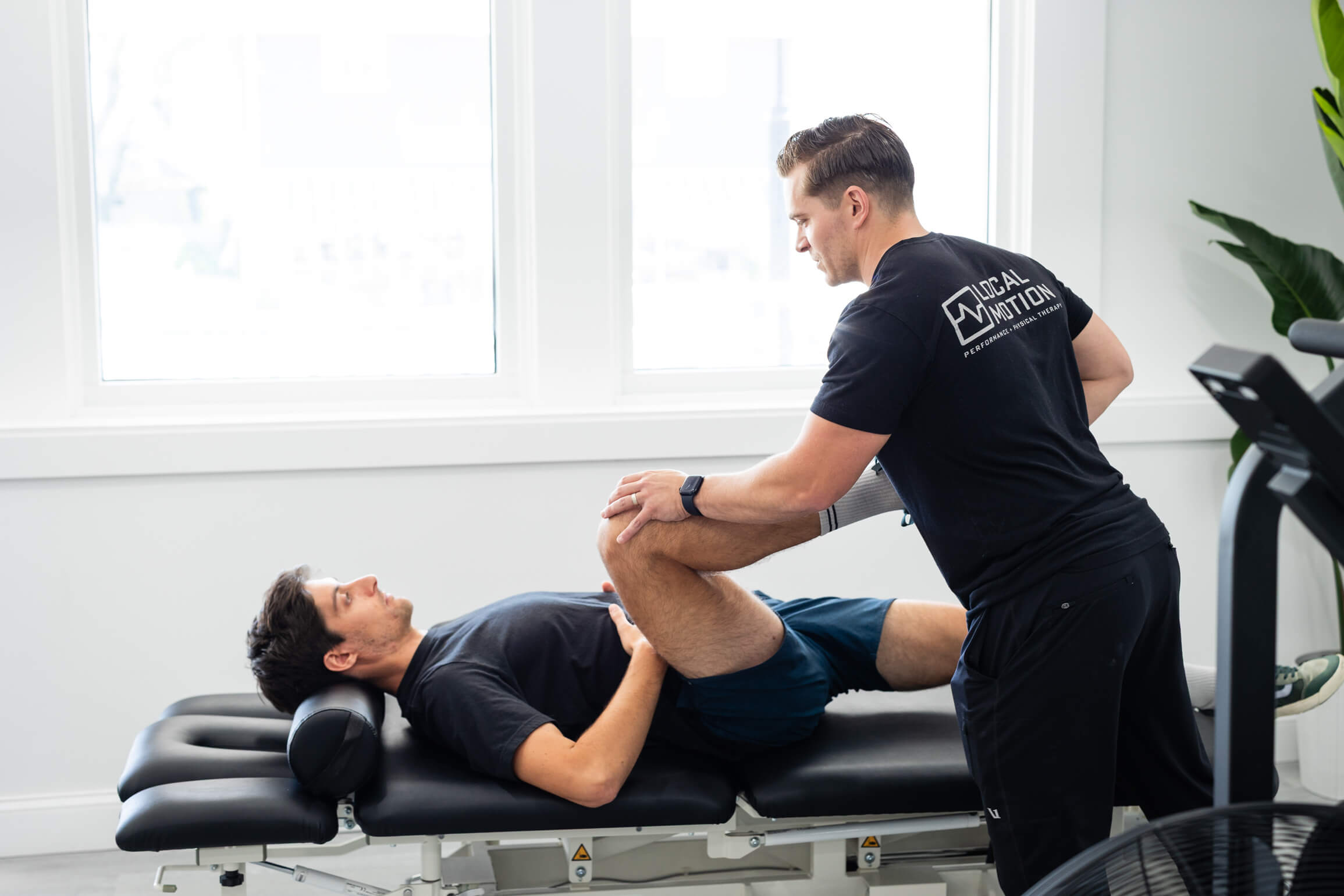 Physical therapist in black Local Motion shirt assists a man lying on a bench by stretching his bent knee towards his chest.