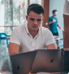 Man in white polo shirt focused on work using a laptop indoors with blurred background.