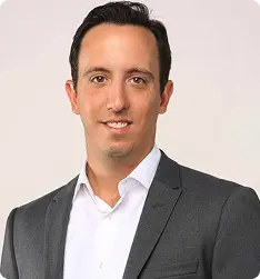 Portrait of a man in a dark suit jacket and white shirt smiling against a plain background.