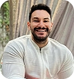 Smiling man with beard wearing a white knit sweater sitting with crossed arms.