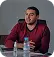Man in a red shirt sitting at a table speaking or presenting with a laptop and microphone.