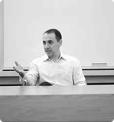Man in white shirt gesturing with hand while sitting behind a desk in a room with a whiteboard.