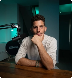 Man with curly hair in white shirt sitting at desk with microphone in a dimly lit room.