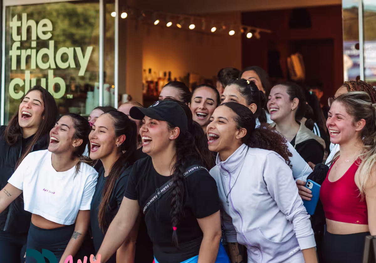 Group of smiling young women standing close together in front of a window with the words 'the friday club' visible.