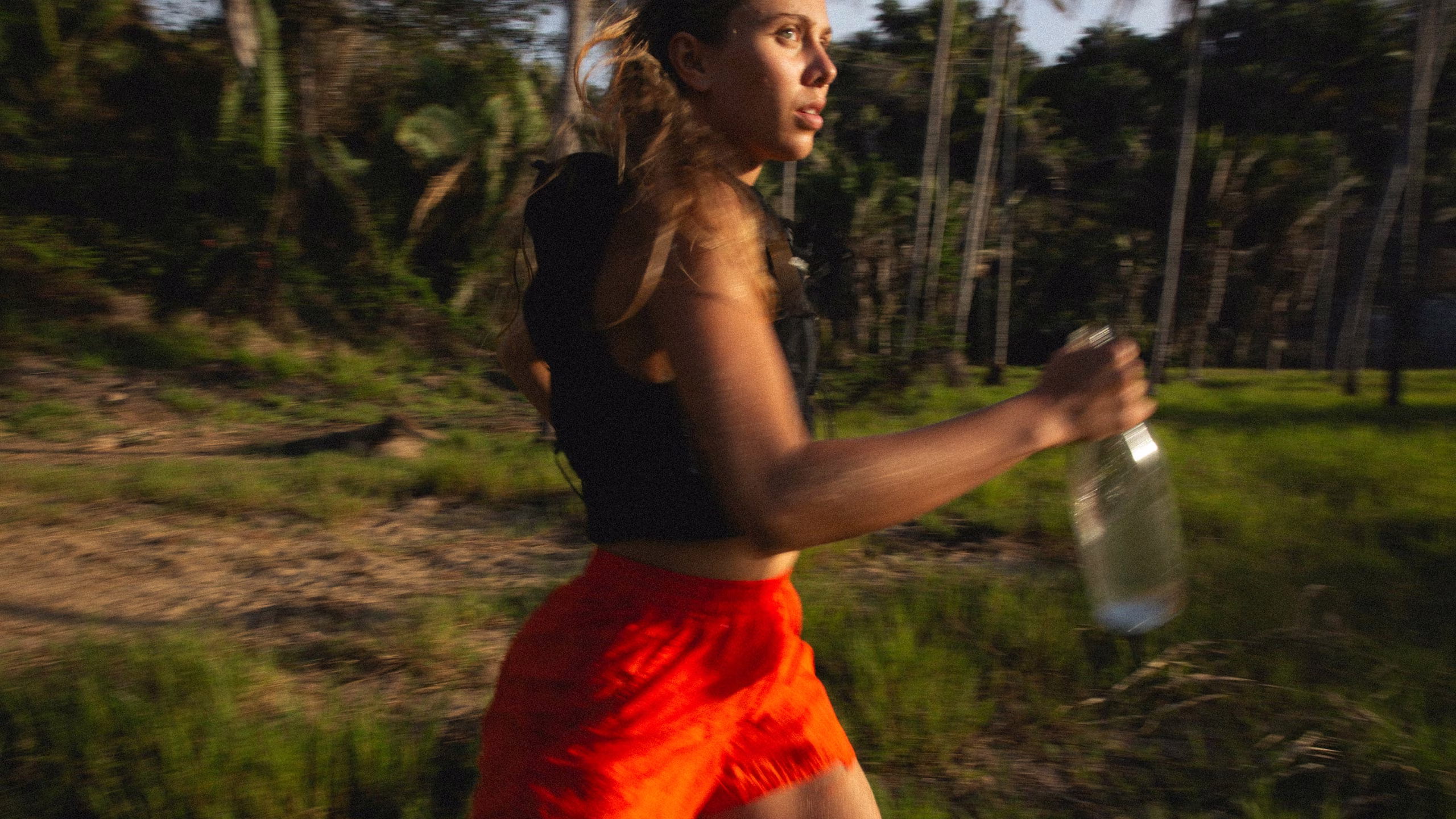 Corredora con shorts rojos atravesando un palmar con una botella transparente de Sip, bajo luz dorada