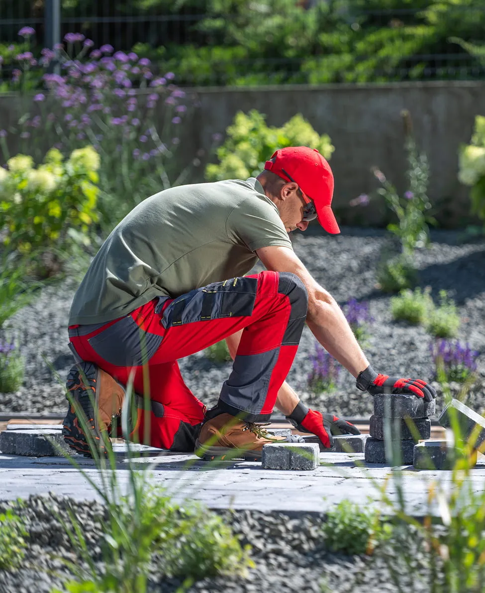 Man in red cap and work gloves kneeling outdoors, arranging rectangular paving stones on the ground.