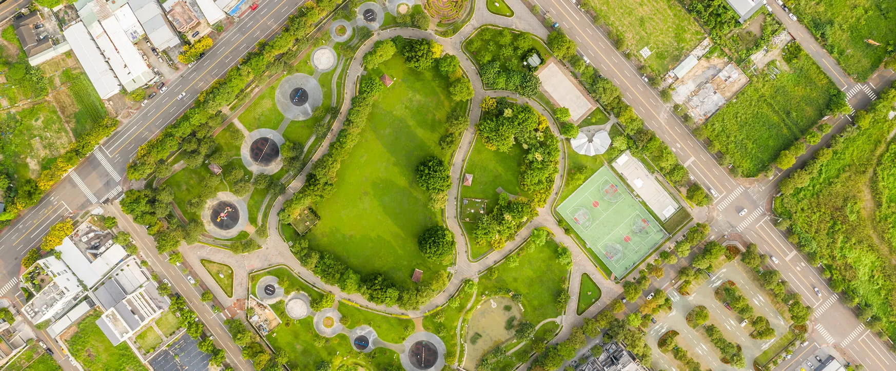 Aerial view of a green park surrounded by roads, featuring walking paths, trees, playgrounds, and a sports court.
