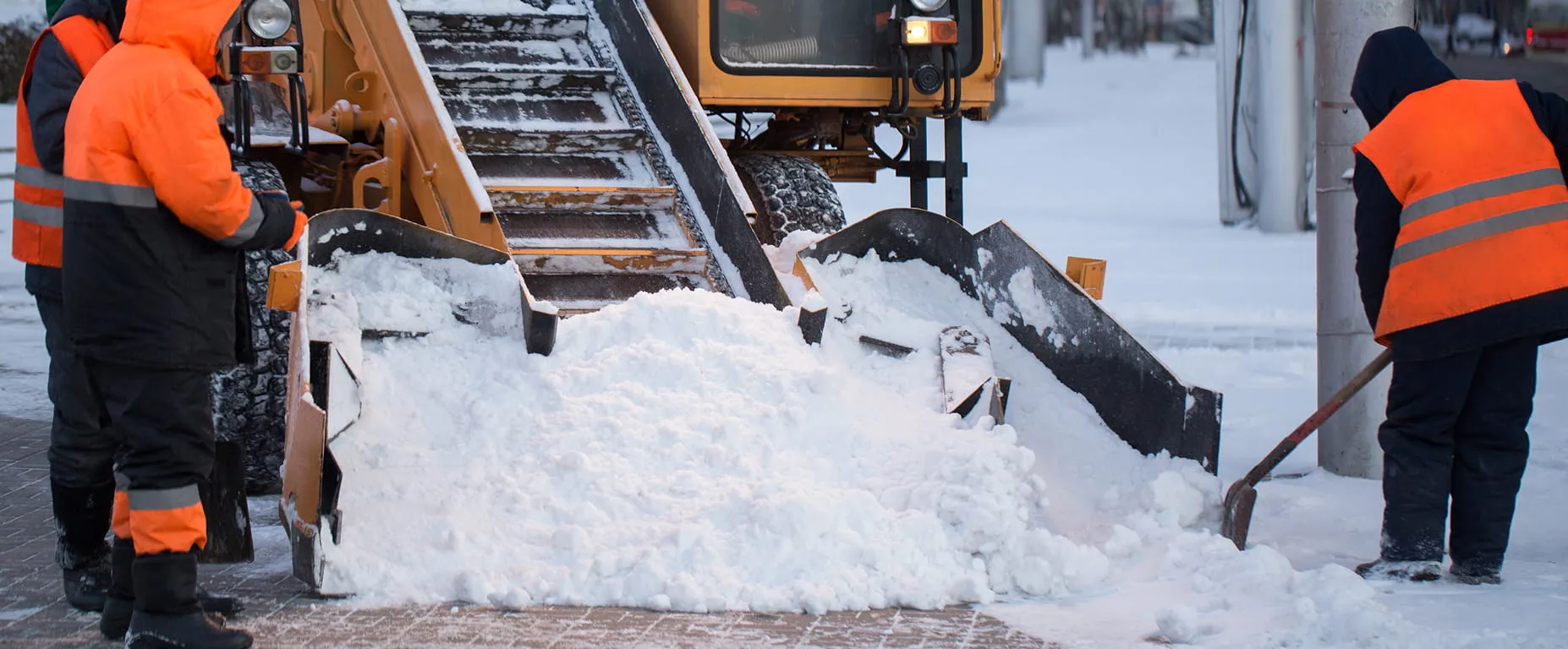 Workers in orange safety jackets clearing snow with a snow removal machine and shovels on a snowy street.