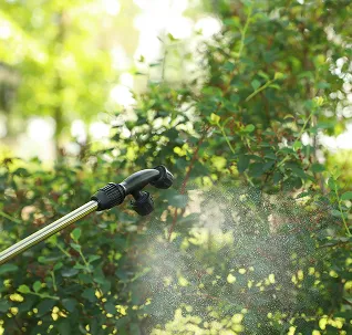 Spray nozzle on a metal wand dispersing mist over green shrubbery outdoors.