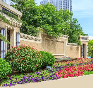 Colorful flower beds with red, purple, and green plants in front of a beige stone wall and lush trees with a tall building in the background.