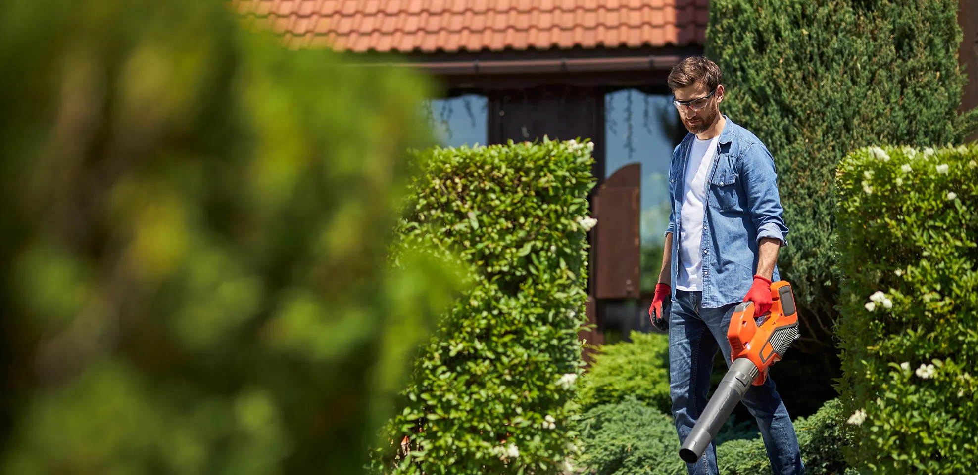 Man wearing safety glasses and red gloves using an orange leaf blower in a garden with trimmed hedges.