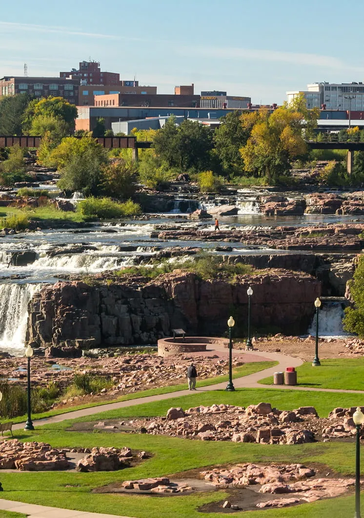 Park with green grass, rocky terrain, and lampposts near cascading waterfalls and a river, with urban buildings visible in the background.