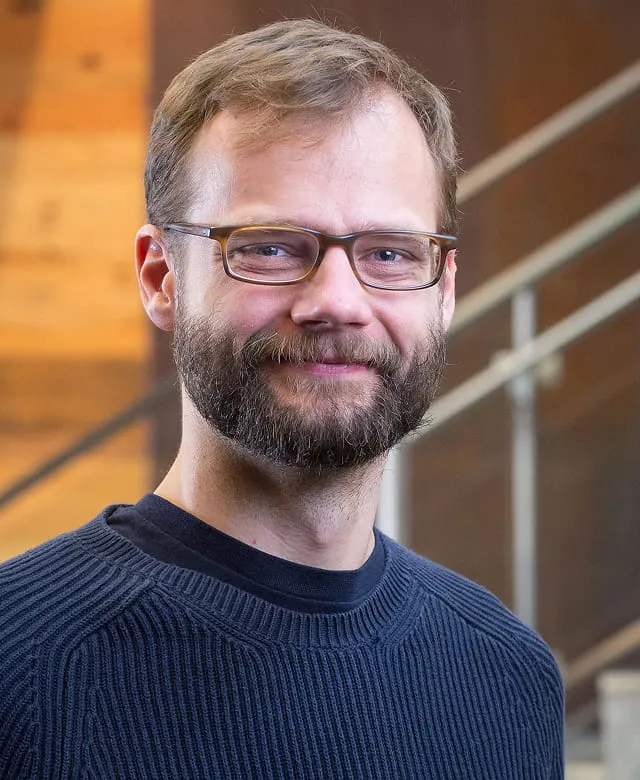 Smiling man with glasses, light brown hair, and beard wearing a dark blue sweater in an indoor setting.