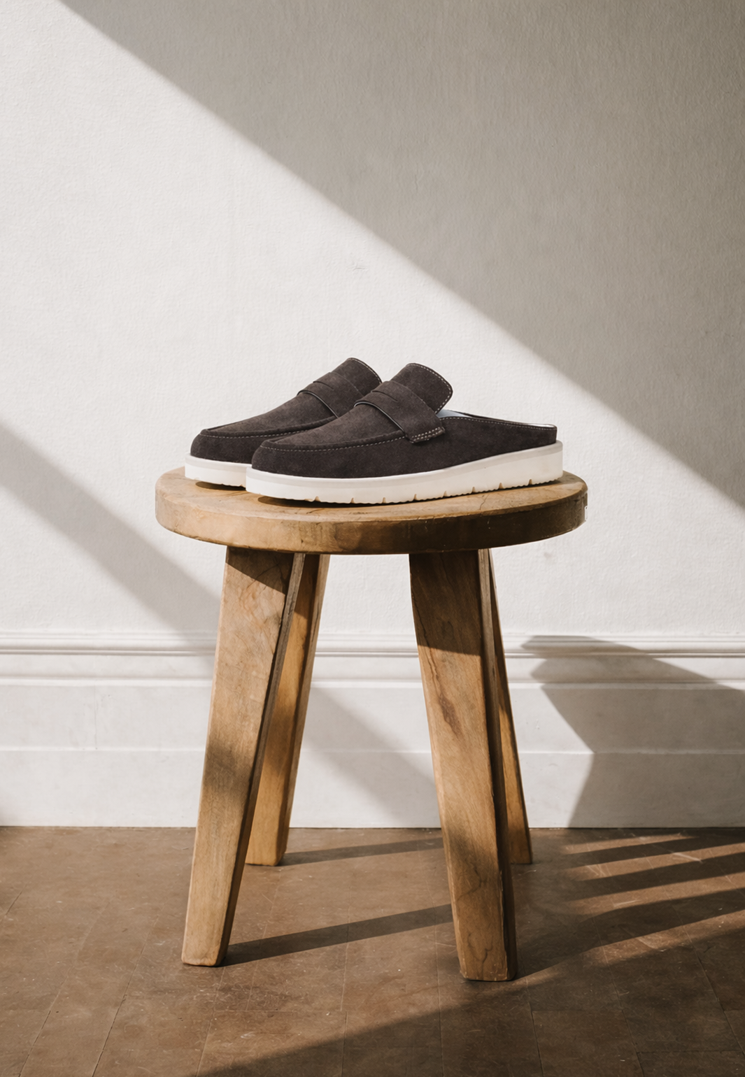 Pair of dark suede KMRO mule slip-on shoes with cream rubber outsoles displayed on a wooden stool.