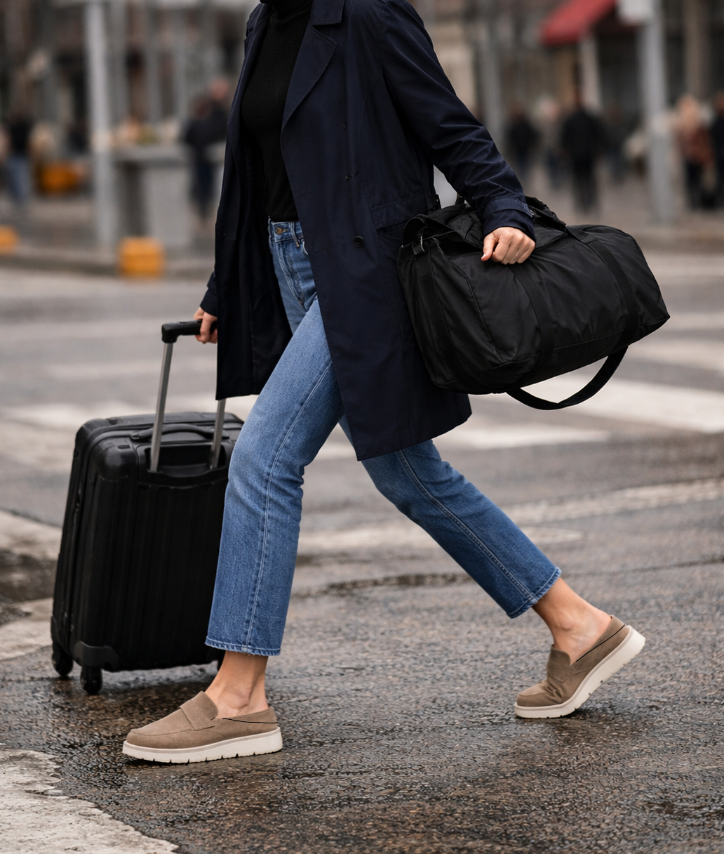 Person walking in suede KMRO mule slip-on shoes while traveling with suitcase and duffel bag in an urban setting.