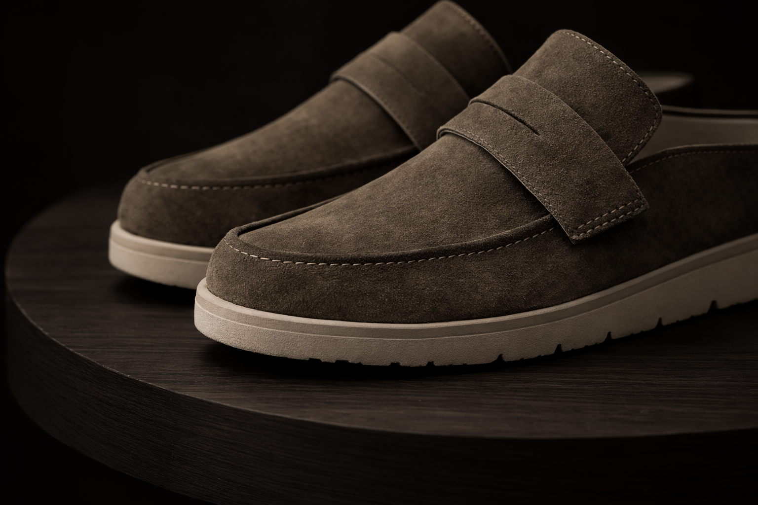A pair of KMRO olive suede mules on a wooden bench in a gallery setting showing side view of their profile.