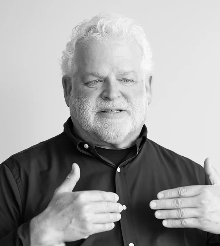 Older man with white curly hair and beard speaking and gesturing with hands near his chest, wearing a dark button-up shirt.