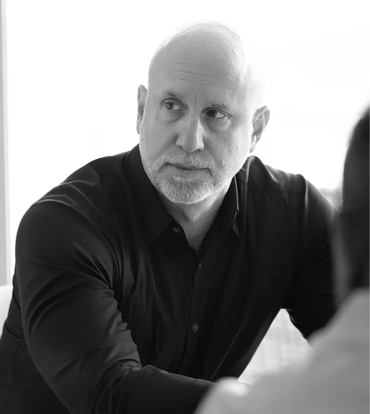 Black and white portrait of a bald man with a beard wearing a black shirt, looking sideways thoughtfully.