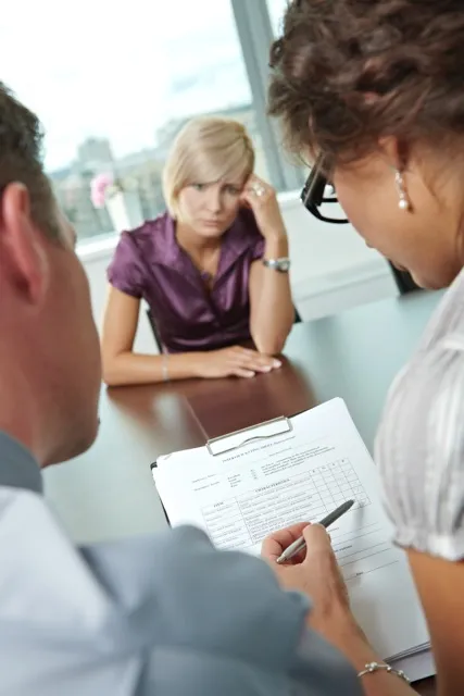 A woman looks stressed while two people review a document on a clipboard during a meeting.