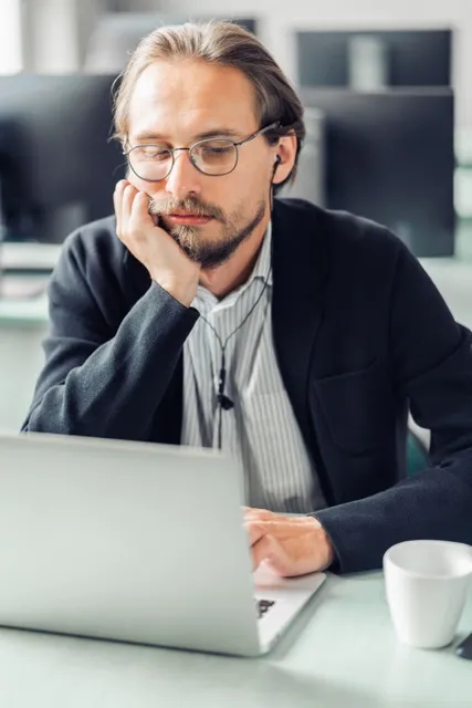 Man with glasses and earphones leaning on hand, working on a laptop at a desk with a white cup nearby.