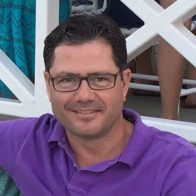 Man with short dark hair and glasses wearing a purple polo shirt, smiling at the camera.