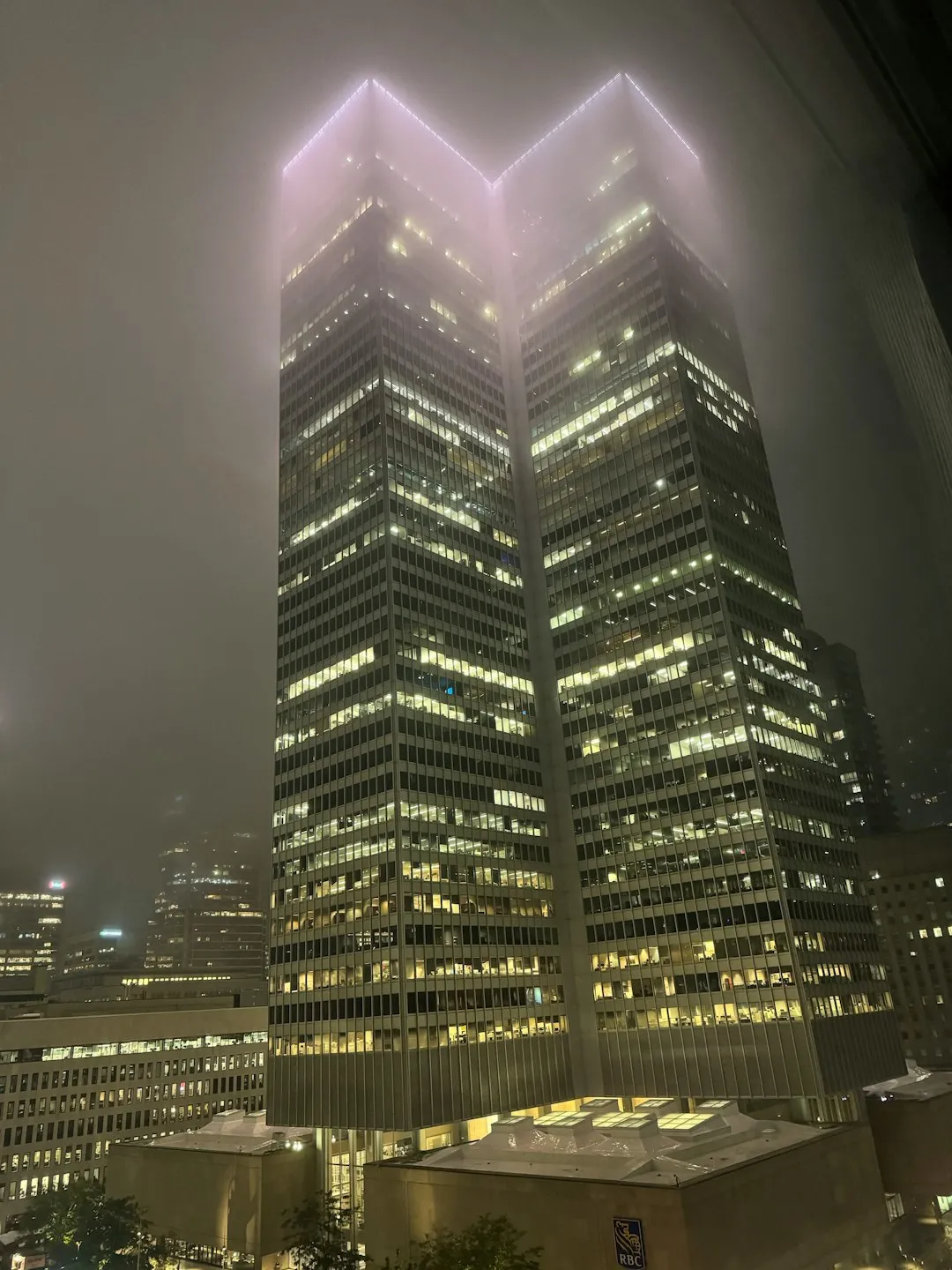 Two illuminated modern skyscrapers at night with fog partially obscuring their tops in a cityscape.