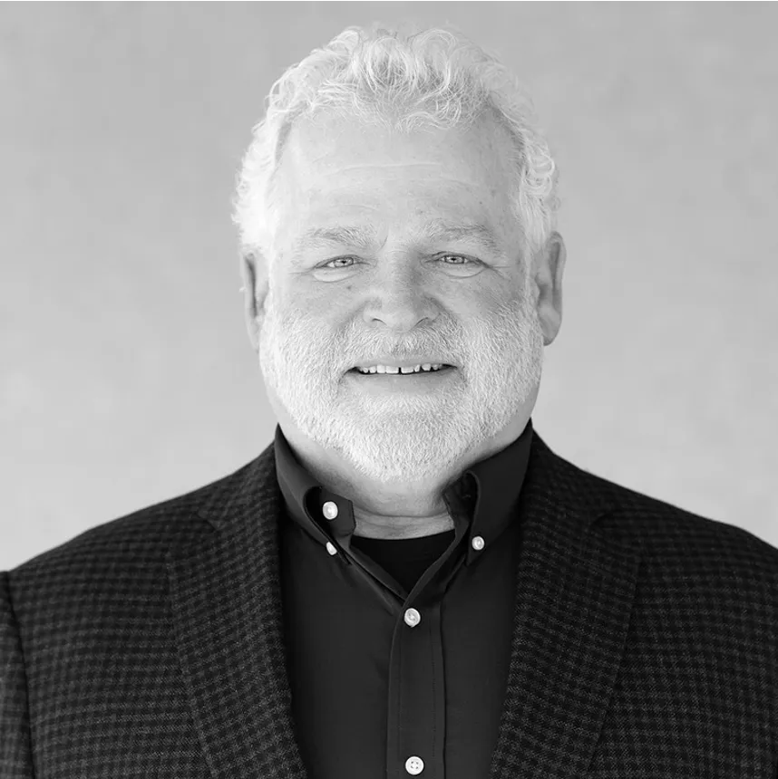 Portrait of a smiling older man with white hair and beard wearing a dark checkered blazer and black shirt.