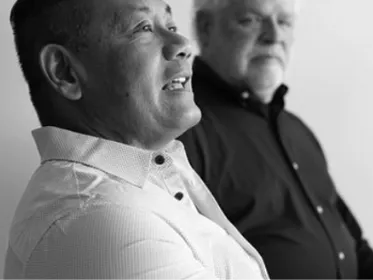 Two elderly men sitting side by side, one speaking, in a black and white portrait.