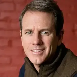 Portrait of a smiling middle-aged man with short brown hair wearing a dark jacket against a red brick wall background.