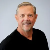 Portrait of a middle-aged man with short gray hair and a goatee, wearing a black shirt and smiling against a light gray background.