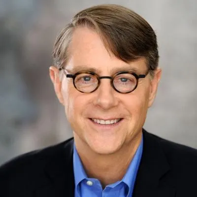 Smiling middle-aged man with short brown hair, wearing round glasses, a blue shirt, and a black blazer.