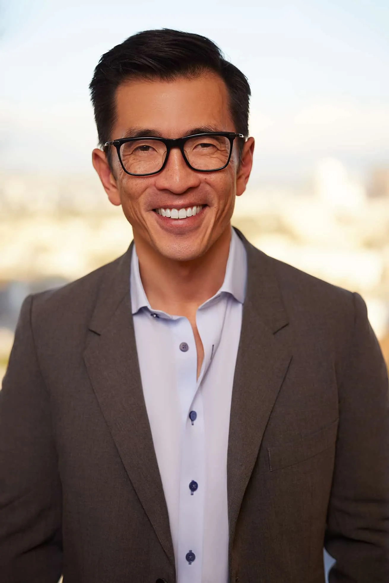 Smiling man with black glasses wearing a brown blazer and light blue shirt standing outdoors.